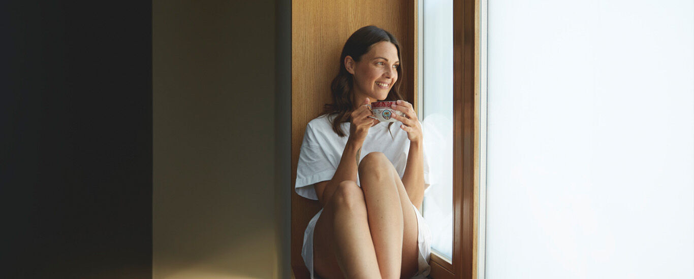Une femme est assise tranquillement pr&egrave;s de la fen&ecirc;tre, en train de boire du th&eacute; ; elle incarne le soin de soi, la s&eacute;r&eacute;nit&eacute; et la sant&eacute; globale des femmes au quotidien
